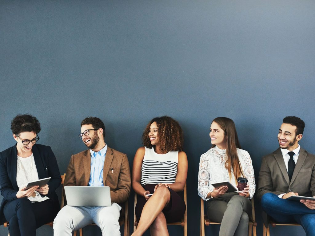 Five smiling adults are sitting in a row on wooden chairs.