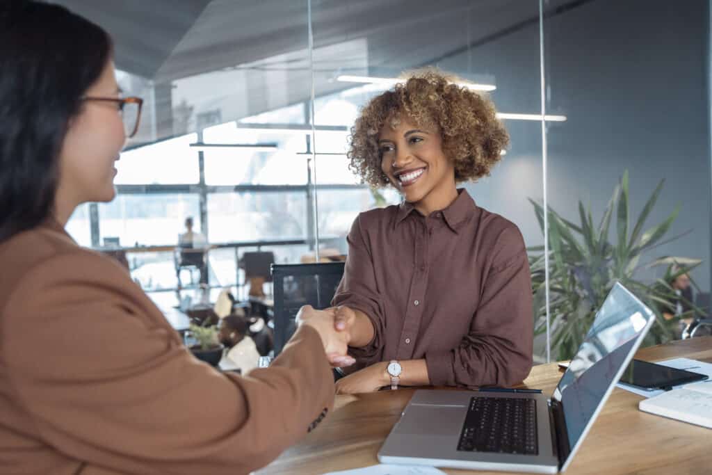 Businesswomenshakehandsatdeskinmodernbusinessofficesmilingatmeetingconveyingagreementwomanpartnershipandprofessionalsuccesspeoplesatisfiedwithresultsofteamworktogether